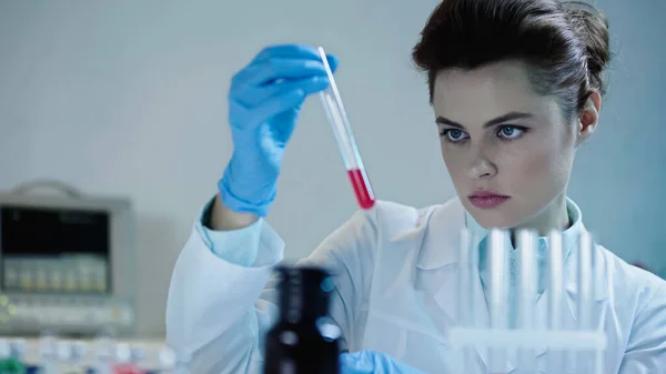 Гарний вчений дивиться на червону рідину в пробірці біля розмитого медичного обладнання — стокове фото