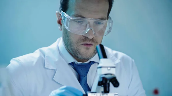 Красивий вчений в захисних окулярах робить аналіз з мікроскопом в лабораторії — стокове фото