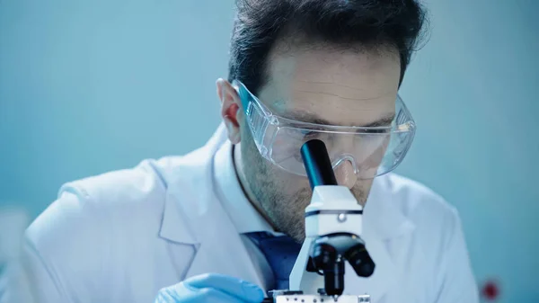 Scientist in goggles and while coat looking through microscope in laboratory — Fotografia de Stock