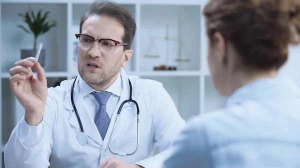 Serious doctor in glasses holding cigarette while warning blurred patient in hospital — стоковое фото