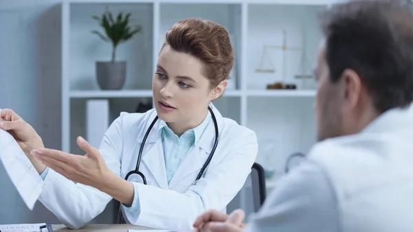 Doctor in white coat pointing with hand at cardiogram and talking to patient in clinic — Fotografia de Stock