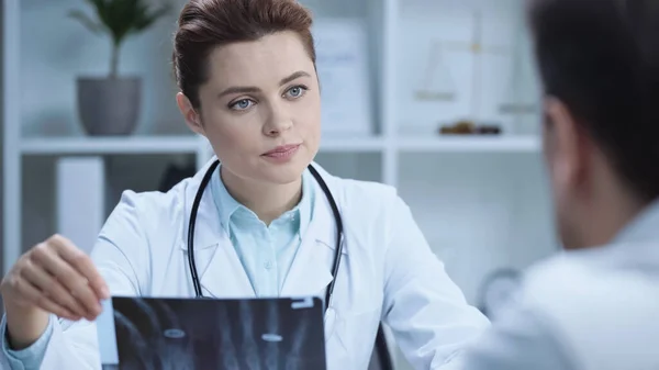 Serious radiologist in white coat holding x-ray scan near man on blurred foreground — Fotografia de Stock