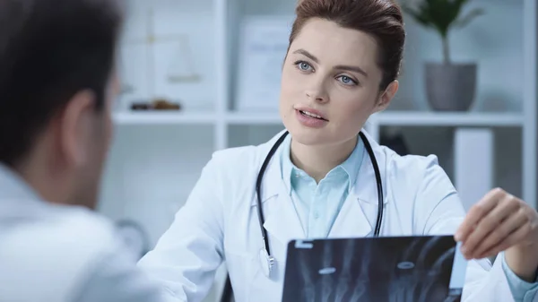 Radiologist in white coat holding x-ray scan and talking with man on blurred foreground — стоковое фото