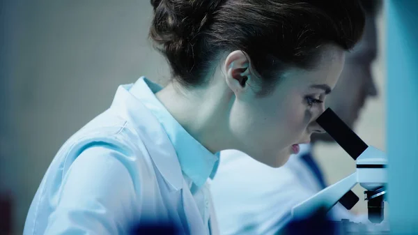 Scientist looking through microscope near blurred colleague in laboratory — Stock Photo