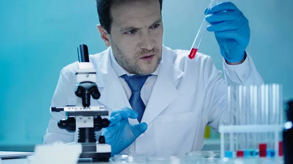 Attentive scientist looking at test tube with red substance near microscope in laboratory — Stock Photo