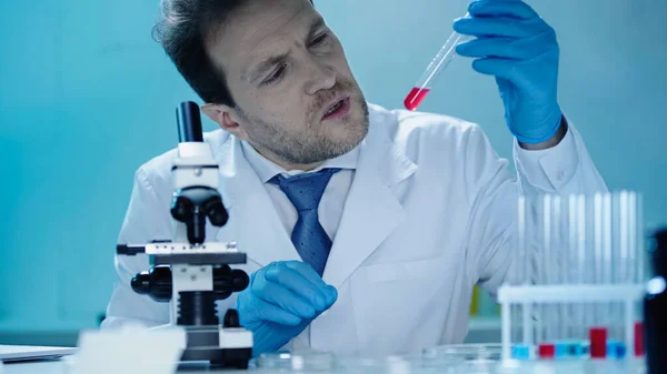 Концентрований вчений, дивлячись на пробірку з червоною речовиною поблизу мікроскопа в лабораторії — стокове фото