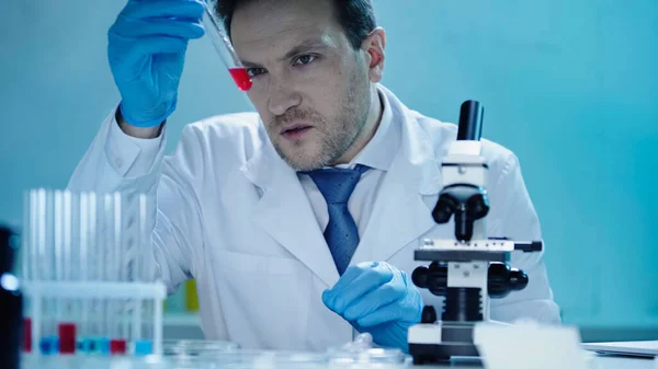 Уважний вчений, дивлячись на пробірку з червоним зразком біля мікроскопа в лабораторії — стокове фото
