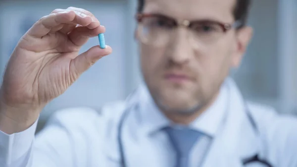 Blurred doctor in glasses and white coat looking at camera and holding capsule — Stock Photo