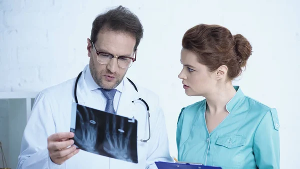 Pretty nurse looking at serious radiologist with x-ray scan in clinic — Stockfoto