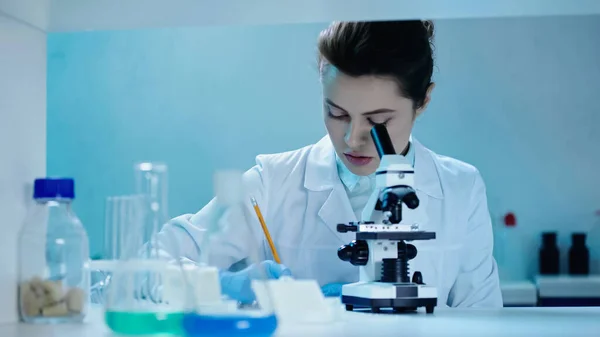 Attractive scientist writing down analysis result near microscope in clinical laboratory — Stock Photo