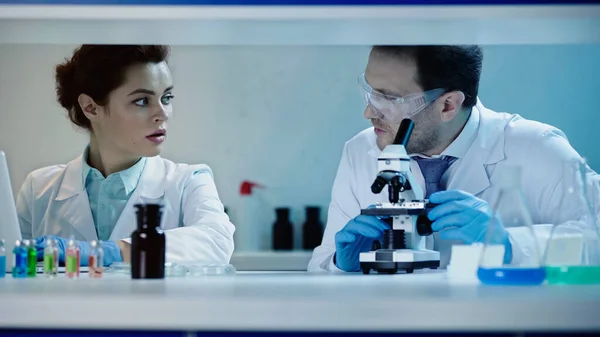 Вчений в окулярах сидить біля мікроскопа і розмовляє з колегою з ноутбуком в лабораторії — стокове фото