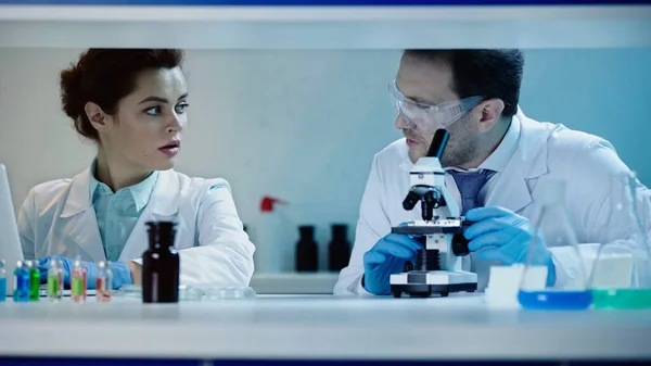Scientist in goggles sitting near microscope and talking to colleague with laptop in laboratory — Stock Photo