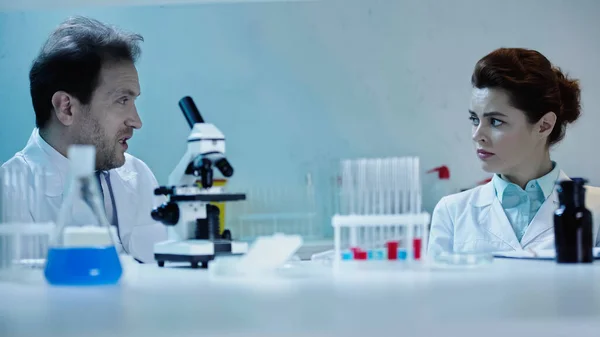 Вчений у білому пальто розмовляє з колегою поблизу мікроскопа в лабораторії — стокове фото