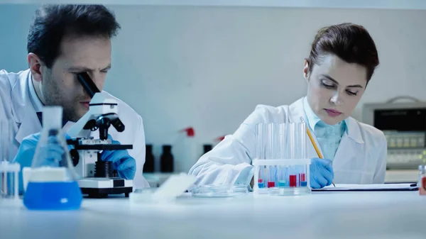 Scientist in white coat looking through microscope near coworker writing result in laboratory — стоковое фото