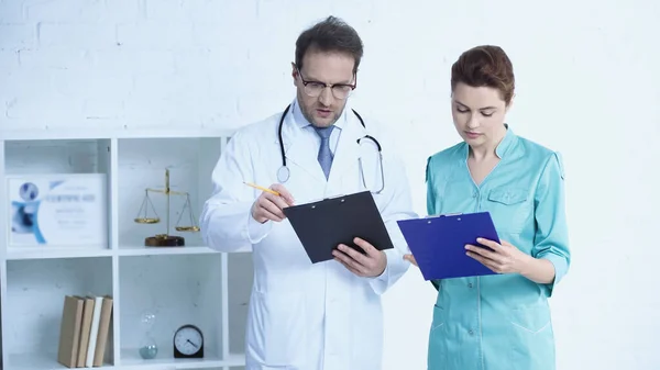 Doctor in glasses and pretty nurse standing with clipboards in clinic — Stock Photo
