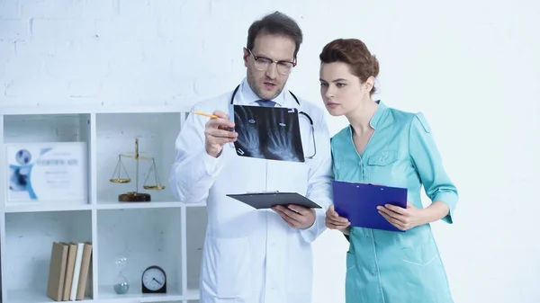 Radiologist in glasses and pretty nurse standing with clipboards and looking at x-ray scan - foto de stock