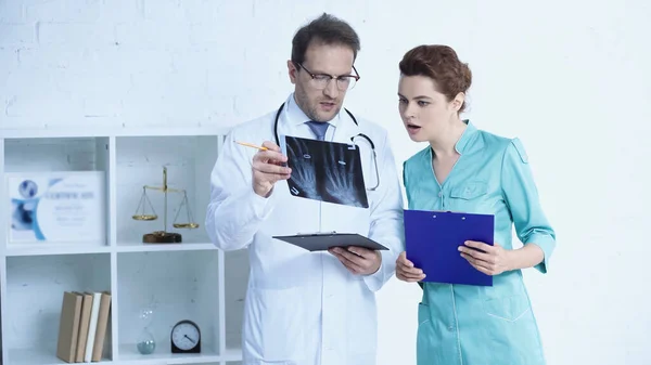 Radiologist in glasses and pretty nurse standing with clipboards and talking while looking at x-ray scan - foto de stock