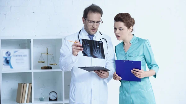 Radiologist in glasses and pretty nurse standing with clipboards and talking while looking at x-ray scan — стоковое фото