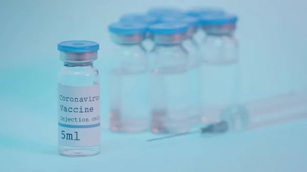 Bottles with coronavirus vaccine lettering near blurred syringe on blue — Foto stock