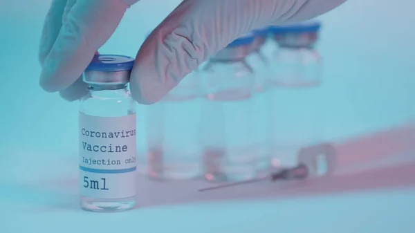 Partial view of scientist in latex glove taking bottle with coronavirus vaccine lettering on blue — Stock Photo