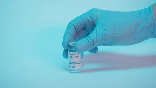 Partial view of scientist in latex glove taking bottle with coronavirus vaccine lettering on blue background — Stock Photo