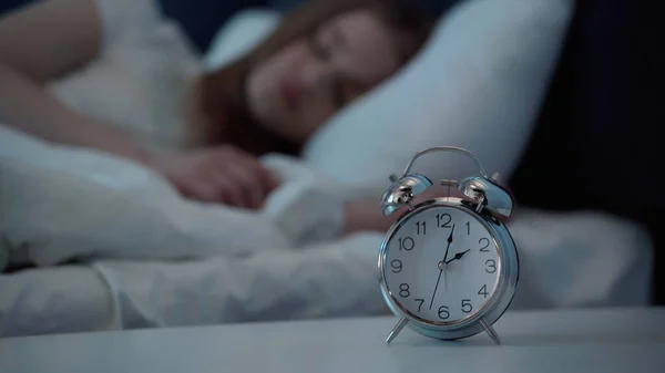 Alarm clock on bedside table near blurred woman sleeping on bed - foto de stock