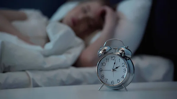 Alarm clock on bedside table near blurred woman lying in bedroom at night - foto de stock