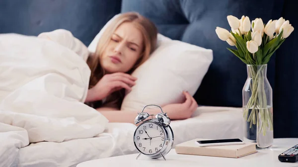 Displeased woman looking at alarm clock near smartphone in bedroom — стоковое фото