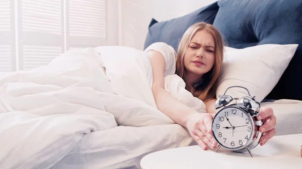Disappointed woman touching alarm clock in bedroom in morning — стоковое фото