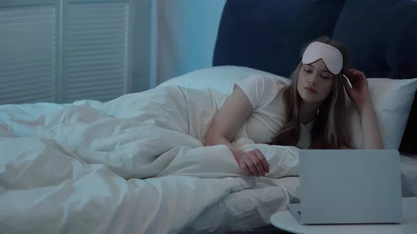 Woman in sleep mask looking at laptop on bedside table in bedroom - foto de stock