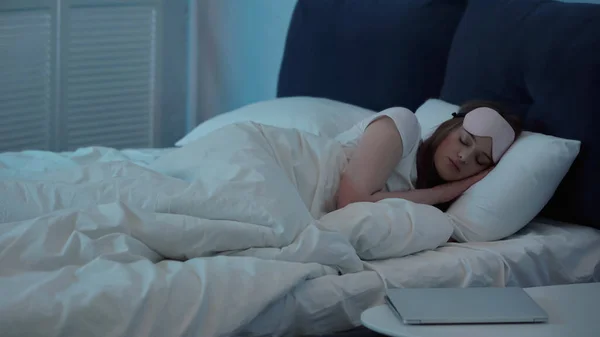 Woman in mask sleeping on bed near laptop on bedside table — Stock Photo
