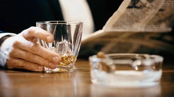 Cropped view of businessman holding glass of whiskey and newspaper near ashtray isolated on black - foto de stock