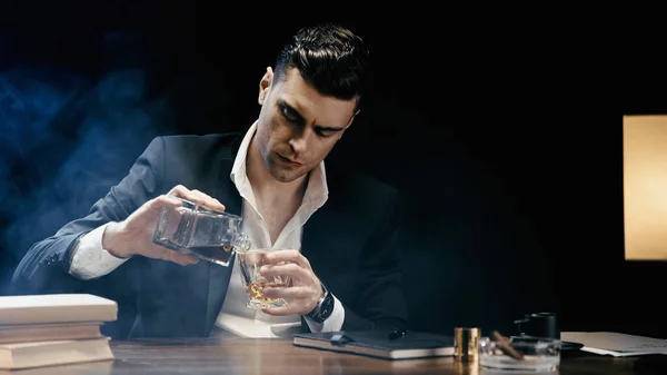 Businessman pouring whiskey near books and cigar on black background with smoke — Photo de stock