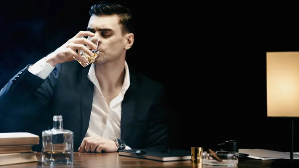 Businessman in suit drinking whiskey near books and ashtray on table isolated on black — Stockfoto