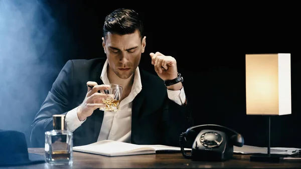 Businessman holding whiskey near notebook and telephone on black background with smoke — Stockfoto