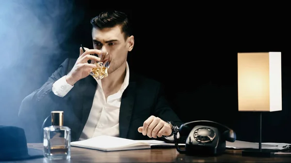 Businessman drinking whiskey near telephone and notebook on black background with smoke — Foto stock
