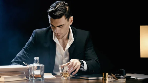 Businessman holding glass of whiskey near books and ashtray on table on black — Photo de stock