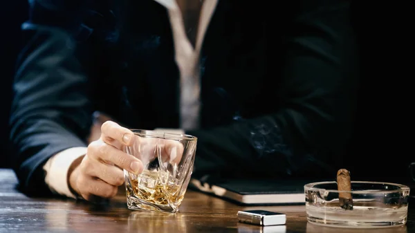 Cropped view of blurred businessman holding glass of whiskey near cigar in ashtray isolated on black — стоковое фото