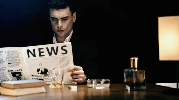 Businessman reading newspaper near whiskey and books on table isolated on black — Photo de stock