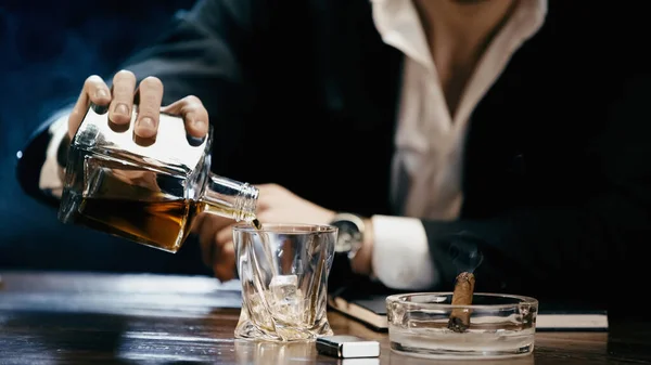 Cropped view of blurred businessman pouring whiskey near cigar in ashtray on black — Fotografia de Stock