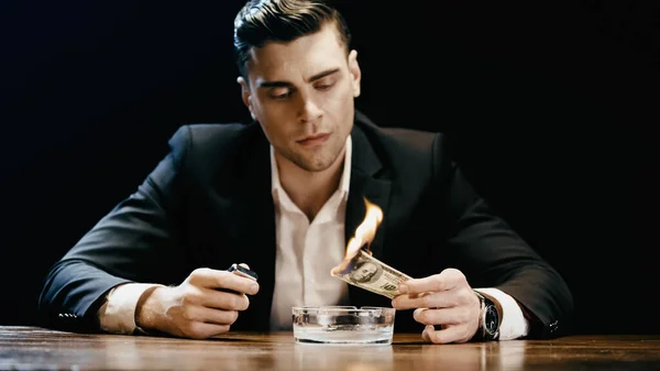 Businessman holding burning dollar and lighter near ashtray on table isolated on black — Stock Photo