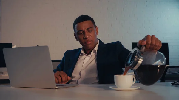 Hombre de negocios afroamericano vertiendo café cerca de la computadora portátil en la oficina por la noche - foto de stock
