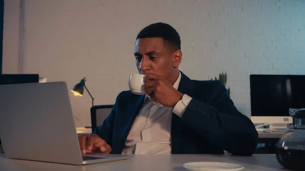 Африканский американский бизнесмен пьет кофе и пользуется ноутбуком в офисе ночью — стоковое фото