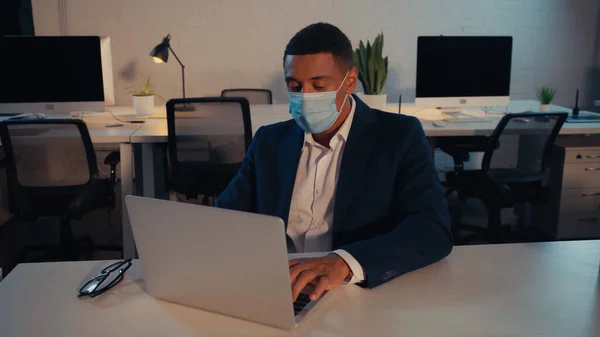 Afroamerikanischer Geschäftsmann in medizinischer Maske mit Laptop nachts im Büro — Stockfoto