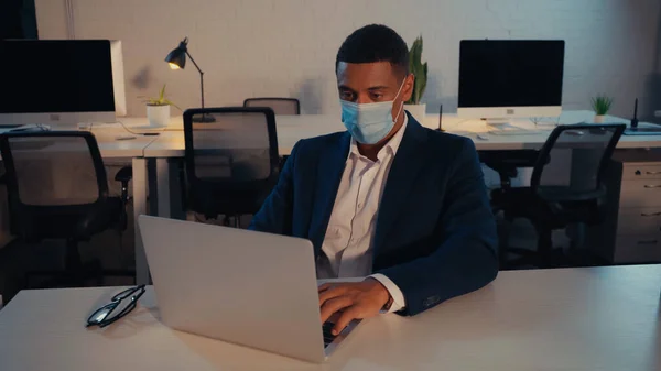 Afroamerikanischer Geschäftsmann in medizinischer Maske mit Laptop in der Nähe von Brillen in der Nacht im Büro — Stockfoto