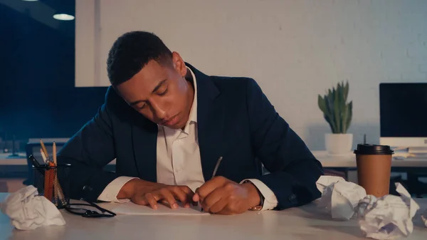 African american businessman writing on paper near crumpled paper and coffee to go in office at night — Stock Photo