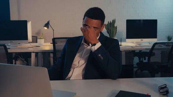 Homme d'affaires afro-américain épuisé assis près d'un ordinateur portable et des lunettes au bureau la nuit — Photo de stock