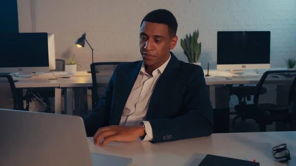 Африканский бизнесмен в костюме смотрит на ноутбук в офисе ночью — стоковое фото
