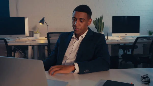 African american businessman in suit looking at laptop in office at night — Stock Photo