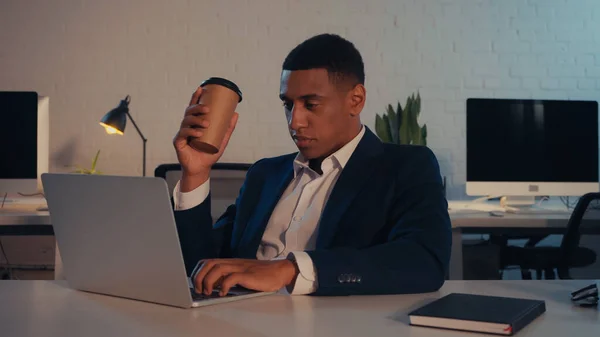 Un homme d'affaires afro-américain tenant un café pour aller et utilisant un ordinateur portable près d'un ordinateur portable au bureau — Photo de stock